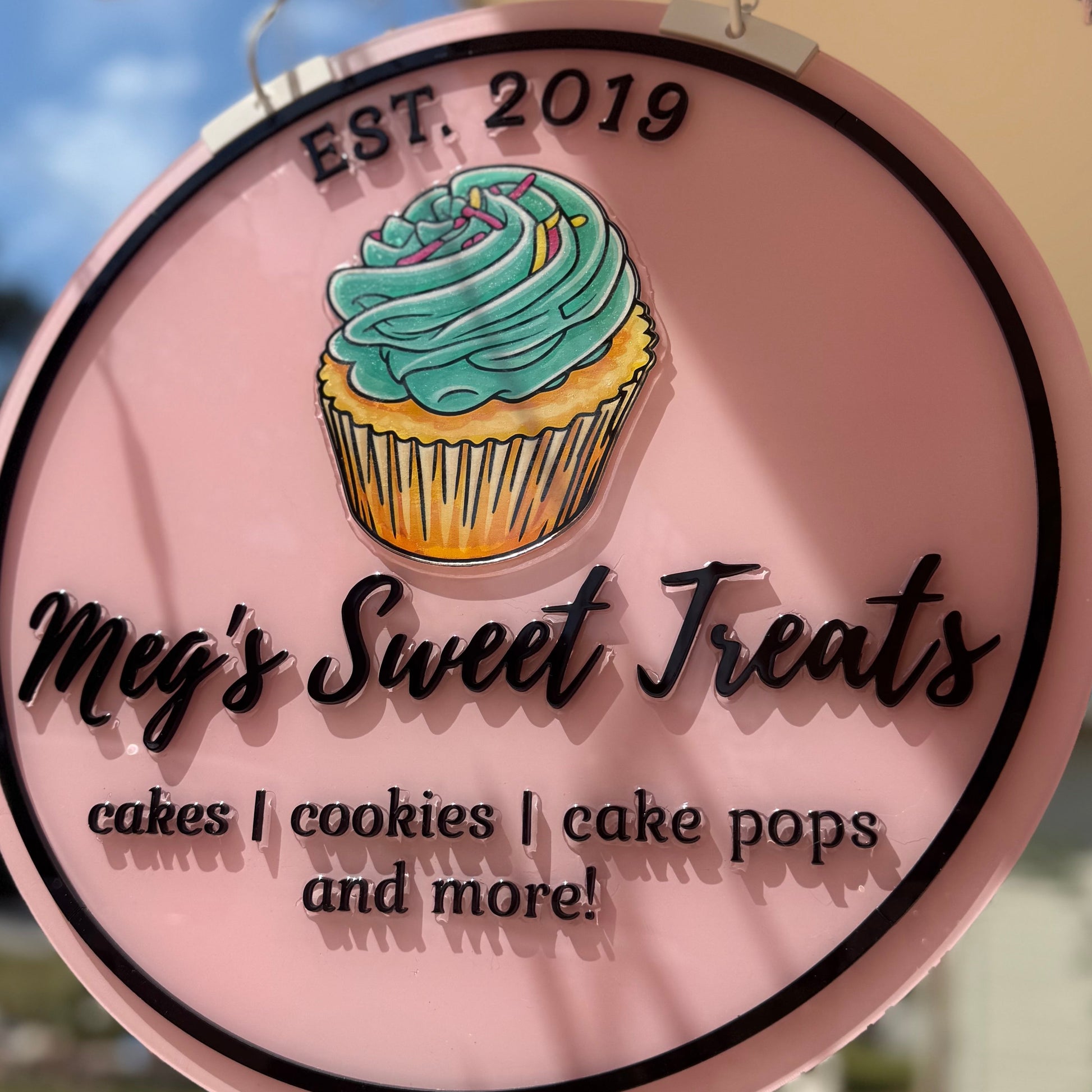 Round sign for Meg's Sweet Treats with a cupcake illustration, hanging outdoors.