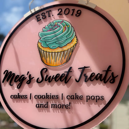 Round sign for Meg's Sweet Treats with a cupcake illustration, hanging outdoors.